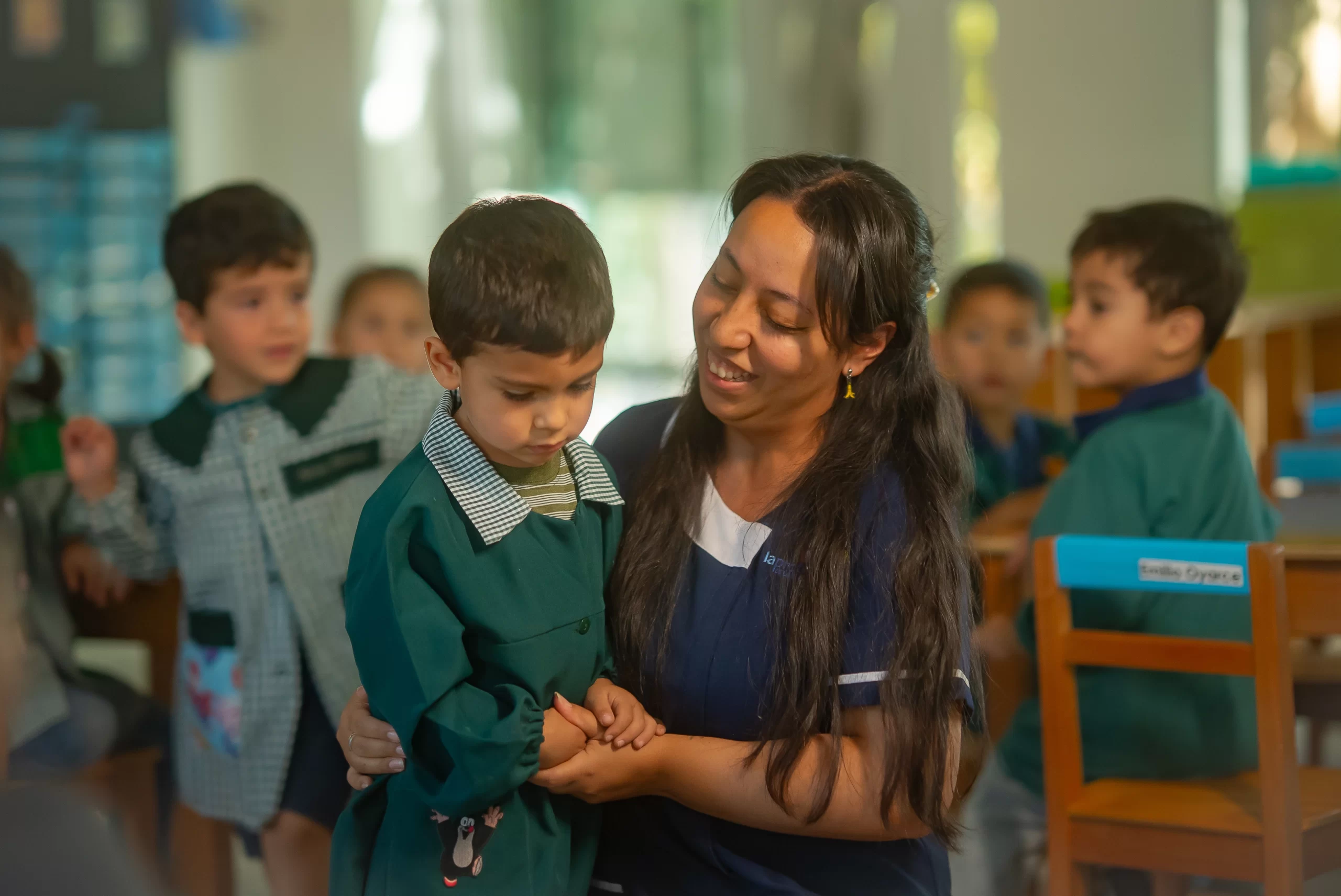 Niño pequeño con uniforme escolar siendo consolado por su profesora en una sala de clases infantil, rodeado de otros niños en un ambiente educativo cálido y seguro.